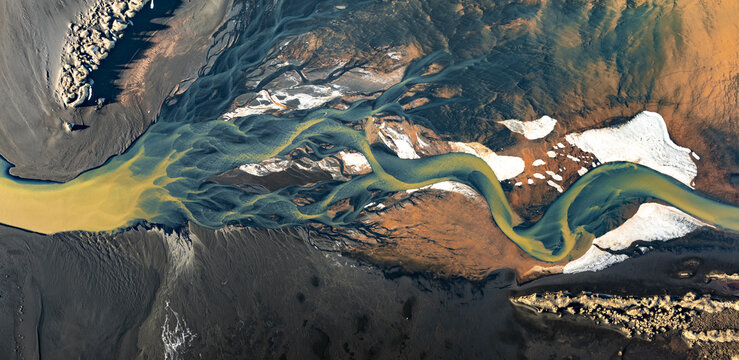 Aerial view of a braided river flowing through dark volcanic sand with sediment-rich water and patches of snow in Iceland.