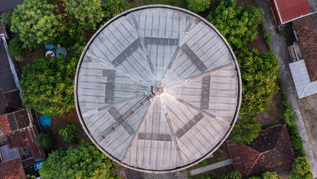 Aerial view of Menara Air Jonggrangan featuring a circular corrugated metal roof surrounded by lush green trees and residential buildings in Kecamatan Klaten Utara, Jawa Tengah, Indonesia.