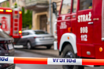 Police warning tape in the foreground, with a blurred fire truck and passenger car parked on a city street in the background, visible "STRAŻ" sign on the red vehicle, emergency services operation. © Adam