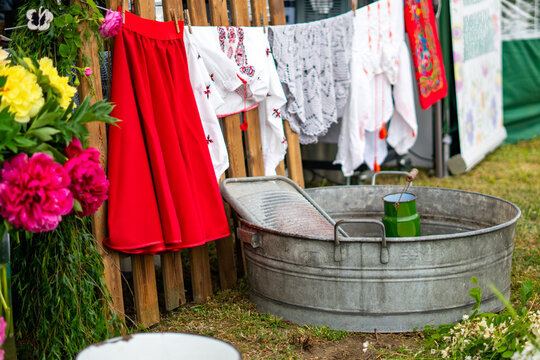 Close-up of a rustic outdoor arrangement featuring a metal washbasin with a washboard, wooden fence with hanging red skirt, traditional white embroidered folk shirts, lace doilies, adorned with vibran