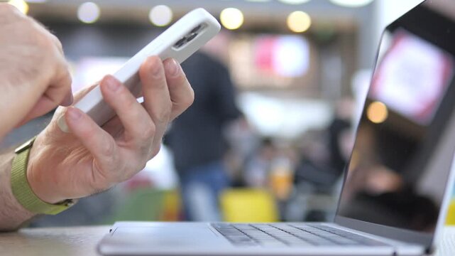 Hands tap phone while laptop sits nearby. Person focused on mobile device. Background shows public space with chairs and people. Suitable for tech, work, or multitasking concepts