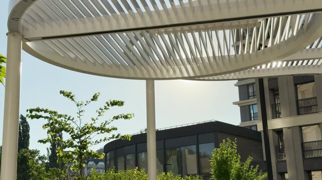A modern white canopy stands in a sunny courtyard in Simferopol, Crimea. Sunlight shines through the slatted roof near tall buildings. This beautiful scene brings a calm, peaceful feeling.