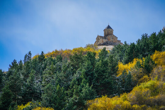 Gergeti Trinity Church, Stepantsminda, Georgia, overlooking a vibrant autumn forest beneath a clear blue sky.
