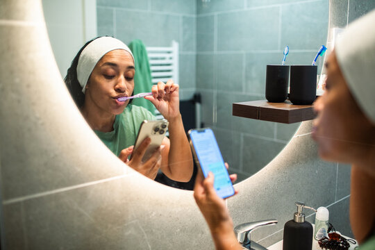 Woman brushing teeth and using smartphone in modern bathroom