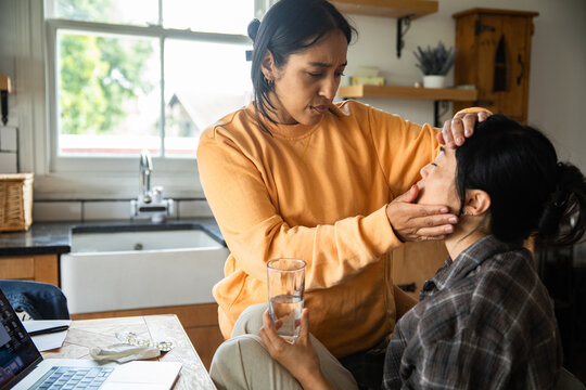 Two women, one checking sick friend's fever in home kitchen