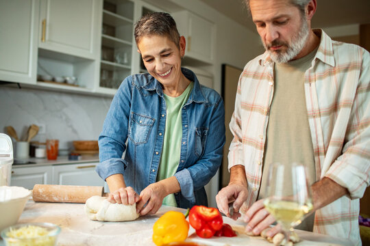 Mature couple kneading dough and chopping vegetables in home kitchen