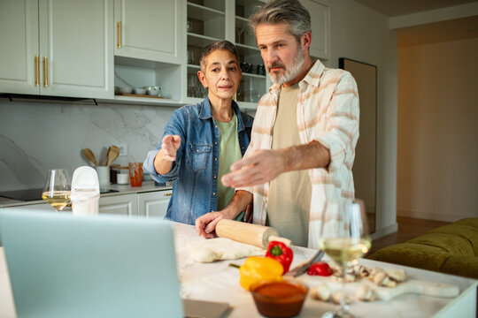 Midlife couple cooking together in home kitchen while following online recipe on laptop