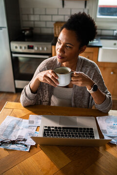 Woman drinking coffee while budgeting with laptop in home kitchen