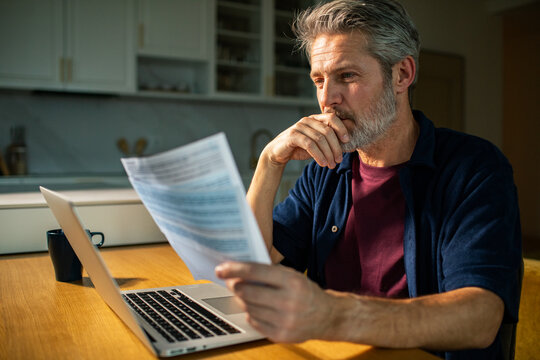 Concerned middle-aged man reviewing bills at kitchen table with laptop
