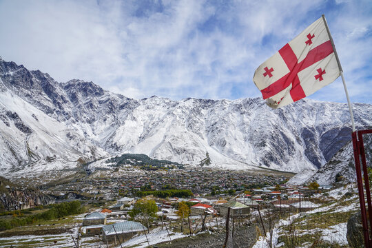 Georgian flag waving over snow-dusted Stepantsminda village against vast Caucasus Mountains under cloudy sky