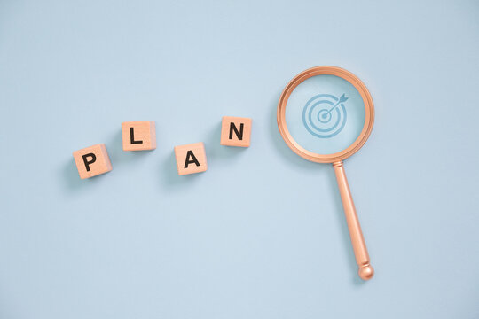 Business planning concept with wooden letter blocks spelling PLAN and magnifying glass highlighting target strategy on blue background, symbolizing goal setting, project management, vision, success.