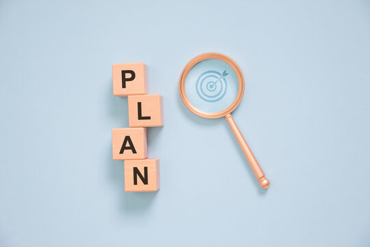 Business planning concept with wooden letter blocks spelling PLAN and magnifying glass highlighting target strategy on blue background, symbolizing goal setting, project management, vision, success.