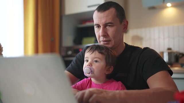 father working from home remotely with baby daughter in his arms. pandemic remote work business concept. father tries to work at home in kitchen, baby fun children interfere sitting on their hands