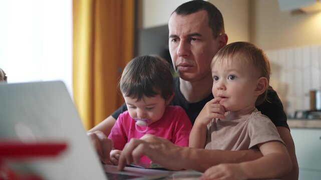 father working from home remotely with two baby in his arms. pandemic remote work business concept. father tries to work at home in kitchen, baby children interfere fun sitting on their hands
