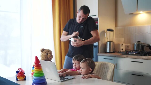 father cooking in kitchen remote work. baby twins playing with laptop in the kitchen. father working at home in the kitchen stirring food in the pan. remote lifestyle work business concept