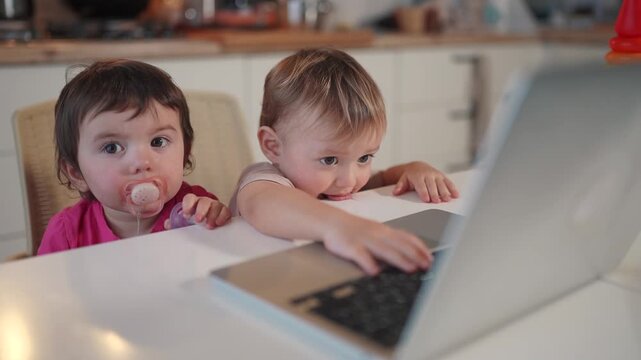 twin baby children playing laptop watching video lifestyle in the kitchen. happy family kid dream concept. baby twins playing video game on laptop looking at screen in kitchen
