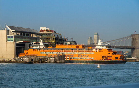 New York, NY - US - Jul 1, 2026 An iconic bright orange Staten Island Ferry vessel rests docked at the Whitehall Terminal in Lower Manhattan, framed by the distant span of the Brooklyn Bridge.