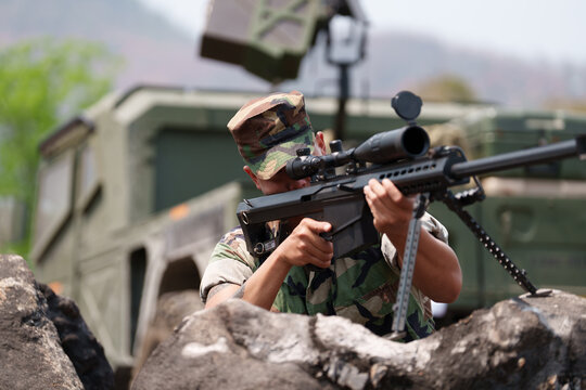 Soldier aims a rifle during a military exercise near armored vehicles in a training area