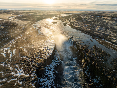 Aerial view of Dettifoss waterfall flowing through a rugged volcanic landscape with mist and golden hour light in Nordurthing, Iceland.