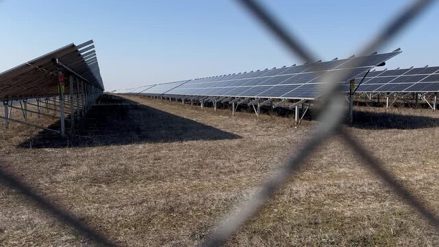 Rack focus through a chain-link security fence reveals rows of tilted solar panels in a dry rural field, warning signs on the steel frames, scorched grass patches under a hazy sky