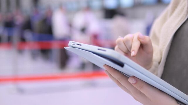 Person holds tablet, taps screen at airport. Background shows blurred travelers and red barriers. Hand focuses on device, fingers interact with touch interface. Scene captures modern travel tech use