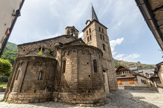 Bossost, Spain. Exterior scenic view of the twelfth century romanesque church featuring its decorated stone apses, a tall bell tower with a slate spire, and traditional houses under a blue sky