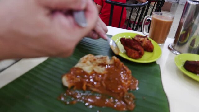 Closeup of person eating roti canai with curry and fried chicken served on banana leaf using fork and spoon in an Indian restaurant in Malaysia