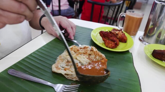Hand with ladle pouring curry onto roti canai served on banana leaf in Indian restaurant in Malaysia. Roti canai is popular breakfast among local Malaysians.