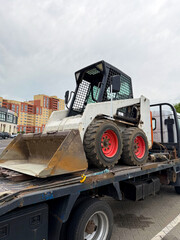 Obraz premium Compact skid steer loader with a bucket attachment parked on a flatbed truck in an urban environment with buildings visible in the background