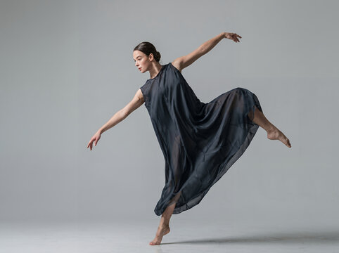 Graceful professional ballerina dancing in a flowing elegant dark grey dress, performing expressive contemporary ballet dance in studio isolated on grey backdrop. Concept of fine art and movement.