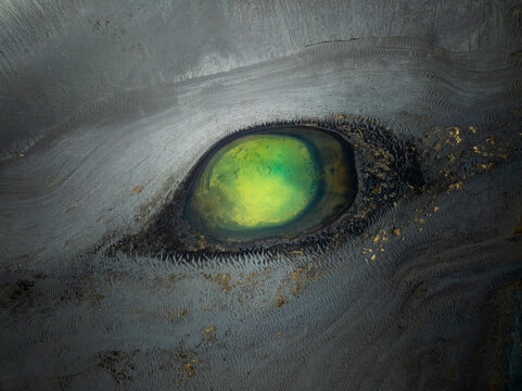 Aerial view of a vibrant green glacial pool surrounded by dark volcanic sand and textured ice formations in Iceland.