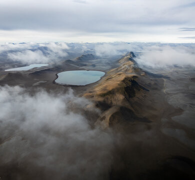 Aerial view of a vast volcanic landscape featuring serene glacial lakes nestled among rugged mountains partially obscured by low-hanging clouds in Iceland.