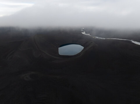 Aerial view of a volcanic crater lake surrounded by dark volcanic terrain and low-hanging clouds in Iceland.