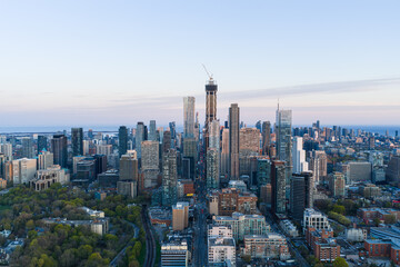 Obraz premium Aerial skyline view over downtown Toronto showing dense modern high rises, central tower under construction, waterfront horizon, parks, streets, and warm evening light