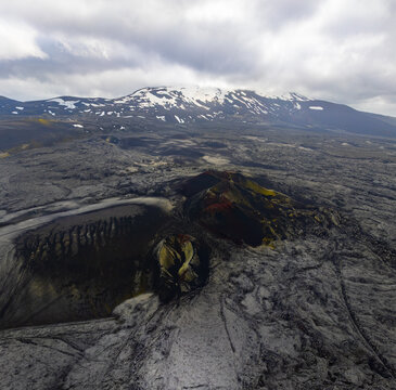 Aerial view of a volcanic crater surrounded by vast solidified lava fields and a snow-capped mountain range under a cloudy sky in Iceland.
