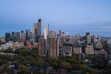 Obraz premium Toronto skyline with downtown high rise towers, CN Tower in distance, evening blue sky, dense urban waterfront district, modern office buildings, apartment blocks, and tree lined foreground