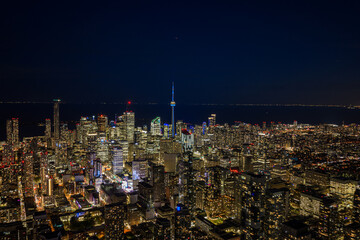 Obraz premium Toronto skyline glowing at night with dense downtown towers, illuminated CN Tower rising above city lights, wide aerial cityscape over Lake Ontario under deep dark sky