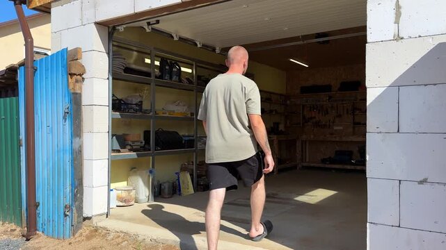 Male technician enters a well-organized garage, walking past shelves filled with tools and equipment, approaching a workbench for maintenance tasks