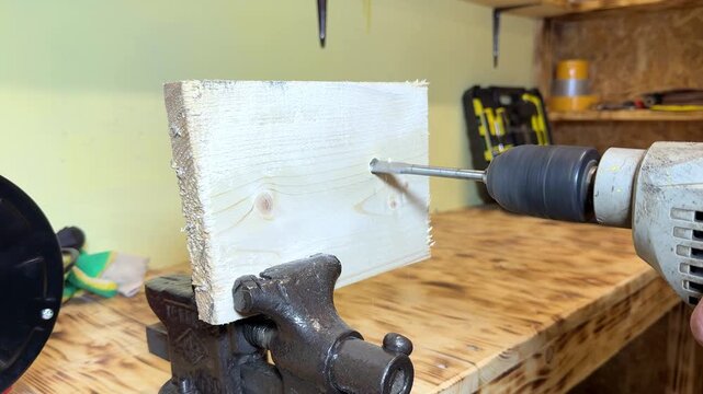 Close-up of a hand using a power drill to create a hole in a wooden plank secured in a vise on a workbench in a workshop setting with tools in the background