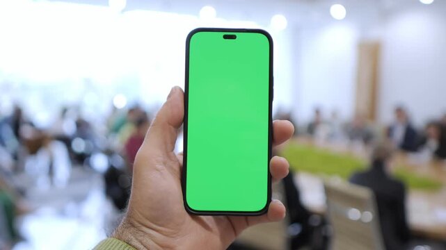 Hand holds smartphone with green screen. Background shows blurred indoor gathering. Focus remains on device for overlays. Ideal for app previews, digital ads, or UI demos