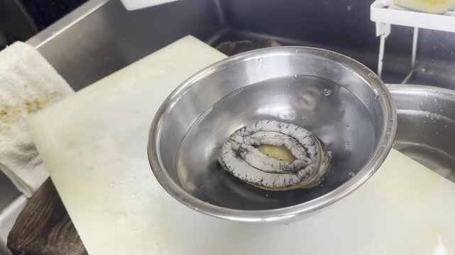 A chef places a cleaned abalone into a bowl of water with kombu kelp, preparing steamed abalone in a Japanese kitchen.