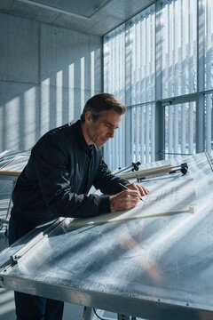Architect leans over a large drafting table to draw precise lines on technical paper with a pencil under strong side sunlight streaming through vertical window slats.