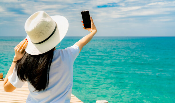 Rear view of a female traveler taking a smartphone selfie on a wooden pier overlooking the turquoise tropical sea during a sunny summer vacation. Travel, social media, adventure, and coastal lifestyle