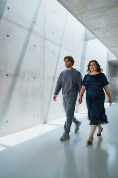 Man and woman walk through a minimalist concrete corridor under bright sunlight hitting the wall at an angle. Motion blur affects their movement as shadows stretch across smooth floor surfaces.