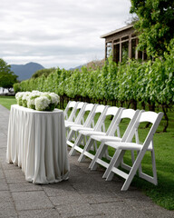 Obraz premium Outdoor wedding venue with white folding chair, floral arrangement, vineyard, green grass, and elegant table setting
