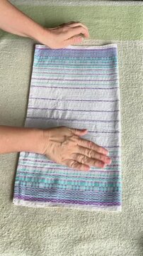 Hands folding a smooth towel after ironing, fabric carefully aligned into square shape, neat preparation for storage, clean and organized, household activity, order and cleanliness. vertical