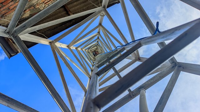 Low-angle perspective looking up inside a galvanized steel lattice tower with a central ladder, creating a striking geometric pattern against a bright blue sky with soft clouds.
