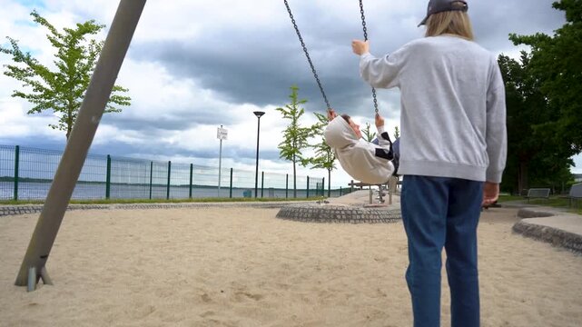 Mother Pushing Child on Swing at Lakeside Playground on Cloudy Summer Day