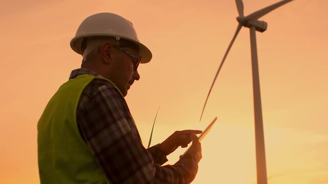 Professional male engineer wearing hard hat and high-visibility jacket stands outdoors wind turbine field. He carefully checks technical information on his tablet, focusing on renewable energy project