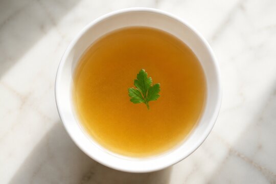 Top-down view of a white bowl with clear broth and a single herb leaf.